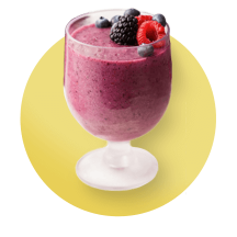 A vibrant purple cherry and mixed berry smoothie topped with fresh blueberries, raspberries, and blackberries in a stemmed glass against a yellow gradient circle.