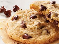 Stack of chocolate chip cookies on brown wax paper, one cookie is broken in half to show warm, chocolatey inside.