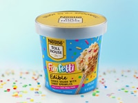 A pint of Funfetti Edible Cookie Dough on a white table, sprinkled with funfetti, against a blue background.