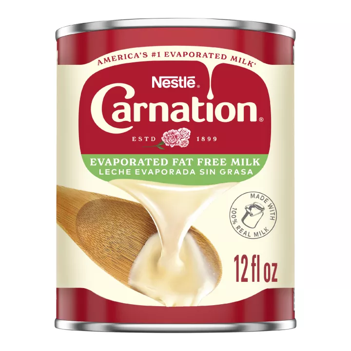 Fotografía frontal de una lata blanca, roja y azul de Leche Evaporada Sin Grasa. Tiene la marca Carnation y el logotipo de la flor, una tapa de botella de plástico azul oscuro, y una imagen de la leche evaporada vertiéndose en una espátula blanca.