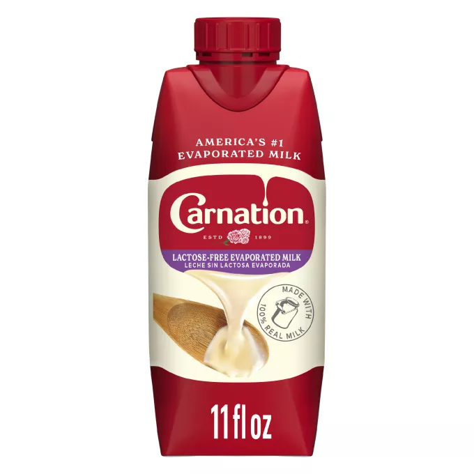 Fotografía frontal de un cartón blanco y rojo octogonal de Leche Evaporada Sin Lactosa. Tiene la marca Carnation y el logotipo de la flor, una tapa de botella de plástico roja y una imagen de leche evaporada vertiéndose sobre una espátula blanca.