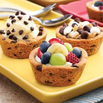 Photo of three cookie-cup desserts on a yellow plate with cream, berries, marshmallows and chocolate chips