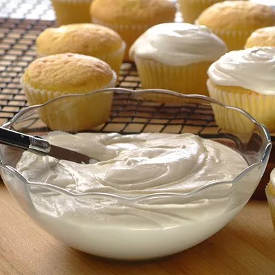 Photograph: bowl of creamy white frosting with spatula; frosted and plain yellow cupcakes on rack