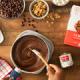 A hand stirs batter in a pot beside a box of Carnation Famous Fudge, a can of Carnation and bowls of chocolate and nuts.